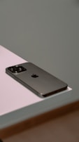 A smartphone with a sleek design and metallic finish rests on the edge of a white surface. The device features a prominent camera array on the back.