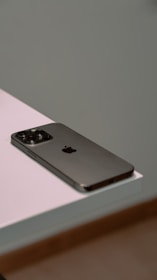 A smartphone with a sleek design and metallic finish rests on the edge of a white surface. The device features a prominent camera array on the back.