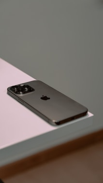 A smartphone with a sleek design and metallic finish rests on the edge of a white surface. The device features a prominent camera array on the back.