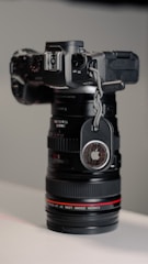 A black professional camera with a zoom lens is positioned vertically, with an Apple AirTag attached to its side using a braided cord. The lens has a subtle red ring near the base, and the background consists of soft, neutral tones.