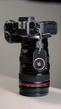 A black professional camera with a zoom lens is positioned vertically, with an Apple AirTag attached to its side using a braided cord. The lens has a subtle red ring near the base, and the background consists of soft, neutral tones.