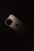 A smartphone with a sleek, metallic finish is placed on a dark surface. The device features a prominent camera module with multiple lenses and an Apple logo in the center.