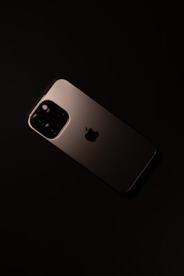 A smartphone with a sleek, metallic finish is placed on a dark surface. The device features a prominent camera module with multiple lenses and an Apple logo in the center.