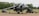 A military aircraft is parked on a tarmac, equipped with various machinery and ground support vehicles positioned nearby. The environment includes an airfield surrounded by trees and open fields, under an overcast sky.