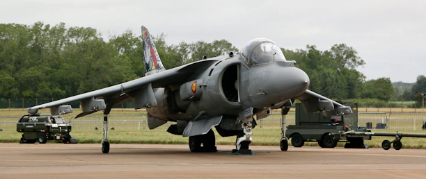 A military aircraft is parked on a tarmac, equipped with various machinery and ground support vehicles positioned nearby. The environment includes an airfield surrounded by trees and open fields, under an overcast sky.