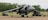 A military aircraft is parked on a tarmac, equipped with various machinery and ground support vehicles positioned nearby. The environment includes an airfield surrounded by trees and open fields, under an overcast sky.