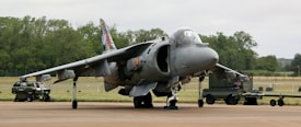A military aircraft is parked on a tarmac, equipped with various machinery and ground support vehicles positioned nearby. The environment includes an airfield surrounded by trees and open fields, under an overcast sky.