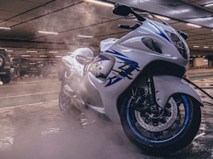 A sleek, white and blue motorcycle parked in an indoor garage with steam or mist surrounding it. Fluorescent lights illuminate the ceiling, and several parked cars are visible in the background.