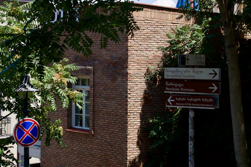 A brick building with a window partially covered by shadows from nearby trees. Signs in both Georgian and English point towards the Botanical Garden and other landmarks. A street lamp is visible beside a no-parking traffic sign.