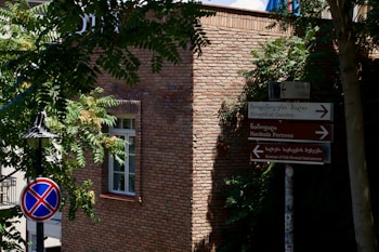 A brick building with a window partially covered by shadows from nearby trees. Signs in both Georgian and English point towards the Botanical Garden and other landmarks. A street lamp is visible beside a no-parking traffic sign.