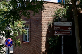 A brick building with a window partially covered by shadows from nearby trees. Signs in both Georgian and English point towards the Botanical Garden and other landmarks. A street lamp is visible beside a no-parking traffic sign.