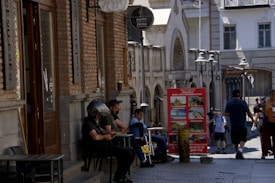 A cobblestone street in an urban setting with people sitting outside a brick building featuring large windows. The building has signs with decorative lettering and a logo. A red informational board displays tour options and stands near the sidewalk, while pedestrians walk nearby. Decorative street lamps line the pathway.
