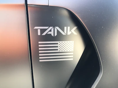 Close-up of a matte black license plate frame engraved with a bold American flag and military insignia, mounted on a sleek car.