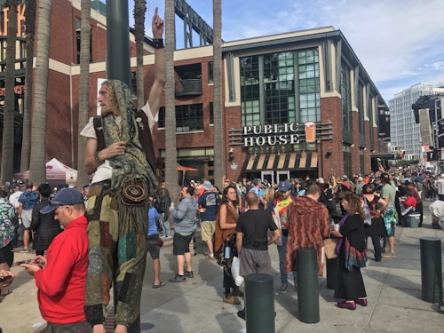 A festive crowd gathers around a modern brick building with a sign that reads 'Public House.' A person dressed in colorful, patterned clothing and scarf stands on an elevated platform, gesturing with one arm raised. Numerous people are mingling on the sidewalk, some wearing vibrant and eclectic attire. Tall palm trees line the area, and the sky is partly cloudy.