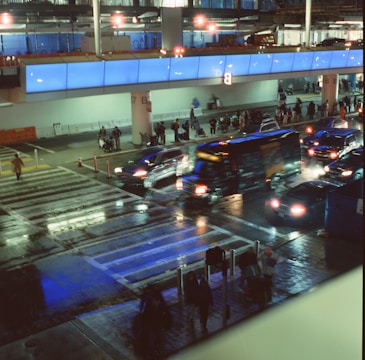 A busy airport terminal area with vehicles halted at a crosswalk. Bright blue lighting illuminates the scene, creating reflections on the wet pavement. People are standing near a building with large windows, and several vehicles, including a bus, are present.