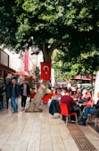 A vibrant street scene with people sitting at outdoor cafe tables. Red chairs and tables are scattered along a tiled sidewalk under a large tree. A Turkish flag is prominently displayed on the tree. Pedestrians walk by, and cars are parked on the street nearby. Lush greenery provides a lively backdrop.