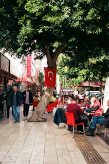 A vibrant street scene with people sitting at outdoor cafe tables. Red chairs and tables are scattered along a tiled sidewalk under a large tree. A Turkish flag is prominently displayed on the tree. Pedestrians walk by, and cars are parked on the street nearby. Lush greenery provides a lively backdrop.