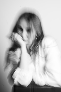 A blurred black and white image of a person sitting with their head resting on their hand. The motion blur creates a dreamy, surreal effect, softening the person’s features and giving the impression of movement or emotion.