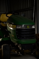 A green and yellow John Deere X320 lawn tractor is parked inside a dimly lit shed. The tractor has a black grille and is partially in shadows. The background includes a metal wall and some indistinct objects.