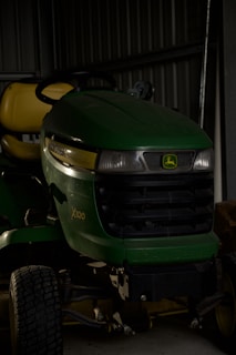 A green and yellow John Deere X320 lawn tractor is parked inside a dimly lit shed. The tractor has a black grille and is partially in shadows. The background includes a metal wall and some indistinct objects.