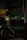 A green and yellow John Deere X320 lawn tractor is parked inside a dimly lit shed. The tractor has a black grille and is partially in shadows. The background includes a metal wall and some indistinct objects.