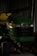 A green and yellow John Deere X320 lawn tractor is parked inside a dimly lit shed. The tractor has a black grille and is partially in shadows. The background includes a metal wall and some indistinct objects.