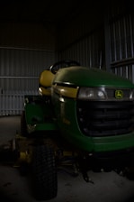 A green and yellow riding lawn mower is parked inside a dimly lit metal shed. The floor is made of concrete, and the shed walls are made of corrugated metal. The brand logo is visible on the front of the mower.