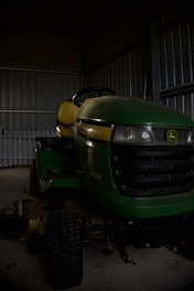 A green and yellow riding lawn mower is parked inside a dimly lit metal shed. The floor is made of concrete, and the shed walls are made of corrugated metal. The brand logo is visible on the front of the mower.