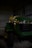 A green and yellow riding lawn mower is parked inside a dimly lit metal shed. The floor is made of concrete, and the shed walls are made of corrugated metal. The brand logo is visible on the front of the mower.