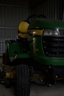 A green riding lawn mower with a yellow seat is parked inside a metal shed. The mower has a large front tire and visible headlights, and part of a white picket fence is visible behind it.