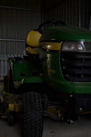 A green riding lawn mower with a yellow seat is parked inside a metal shed. The mower has a large front tire and visible headlights, and part of a white picket fence is visible behind it.