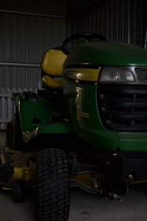 A green riding lawn mower with a yellow seat is parked inside a metal shed. The mower has a large front tire and visible headlights, and part of a white picket fence is visible behind it.