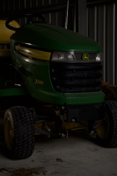 A green John Deere X320 lawn tractor is parked inside a dimly lit garage or shed. The tractor has yellow accents and features large black tires. The setting appears industrial with metal walls in the background.
