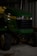 A green John Deere X320 lawn tractor is parked inside a dimly lit garage or shed. The tractor has yellow accents and features large black tires. The setting appears industrial with metal walls in the background.
