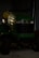 A green John Deere riding lawn mower is parked inside a dimly lit metal garage. The mower features a yellow seat and black steering wheel. Its front grille is dark, casting shadows, and the surface reflects some light.