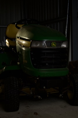 A green John Deere riding lawn mower is parked inside a dimly lit metal garage. The mower features a yellow seat and black steering wheel. Its front grille is dark, casting shadows, and the surface reflects some light.