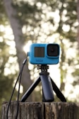 A blue action camera is mounted on a black tripod, placed on a tree stump in an outdoor setting. The background features a soft bokeh effect with warm tones, suggesting sunlight filtering through trees. The camera has a compact build with a lens and display screen.
