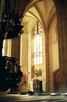 A grand, Gothic-style interior features towering columns and intricate architectural details. The light filters through a tall, stained glass window, casting colorful patterns on the stone walls. Ornate wooden carvings adorn a prominent pulpit or lectern, adding to the space's historical and spiritual atmosphere.