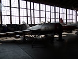 A vintage military aircraft is parked inside a dimly lit hangar with large grid-like windows casting light patterns on the floor. The aircraft has the number 23 on its side and features a star emblem, typical of Soviet-era designs. Another aircraft is partially visible in the background.