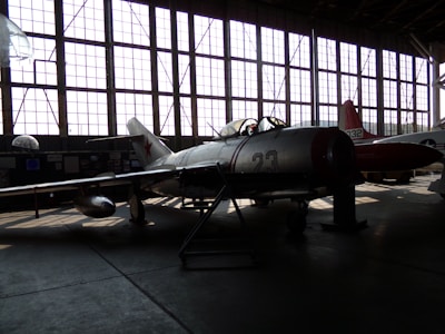 A vintage military aircraft is parked inside a dimly lit hangar with large grid-like windows casting light patterns on the floor. The aircraft has the number 23 on its side and features a star emblem, typical of Soviet-era designs. Another aircraft is partially visible in the background.