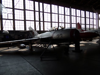 A vintage military aircraft is parked inside a dimly lit hangar with large grid-like windows casting light patterns on the floor. The aircraft has the number 23 on its side and features a star emblem, typical of Soviet-era designs. Another aircraft is partially visible in the background.