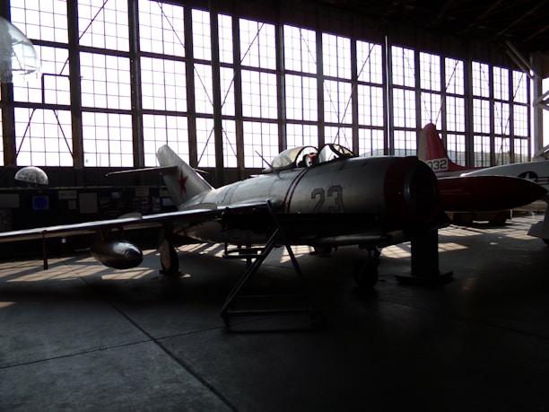 A vintage military aircraft is parked inside a dimly lit hangar with large grid-like windows casting light patterns on the floor. The aircraft has the number 23 on its side and features a star emblem, typical of Soviet-era designs. Another aircraft is partially visible in the background.