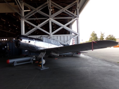 A vintage silver aircraft is positioned inside a hangar, partially outside with its wings stretching into the sunlit area. The aircraft is parked on a concrete floor with visible support structures of the hangar in the background. Trees can be seen through the open hangar doors.