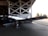A vintage silver aircraft is positioned inside a hangar, partially outside with its wings stretching into the sunlit area. The aircraft is parked on a concrete floor with visible support structures of the hangar in the background. Trees can be seen through the open hangar doors.