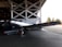 A vintage silver aircraft is positioned inside a hangar, partially outside with its wings stretching into the sunlit area. The aircraft is parked on a concrete floor with visible support structures of the hangar in the background. Trees can be seen through the open hangar doors.