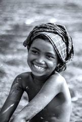 A young child with a big smile is seen in this black-and-white photo. The child is shirtless and has paint smeared on their arms and body. They are wearing a plaid cloth wrapped around their head. The background appears blurred, focusing attention on the child's joyful expression.