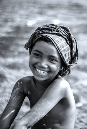 A young child with a big smile is seen in this black-and-white photo. The child is shirtless and has paint smeared on their arms and body. They are wearing a plaid cloth wrapped around their head. The background appears blurred, focusing attention on the child's joyful expression.