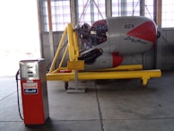 An old gasoline pump with the brand 'Good Gulf' stands next to a disassembled aircraft engine display labeled 'Lady Annie'. The engine is mounted on a yellow stand inside an industrial hangar with large windows.