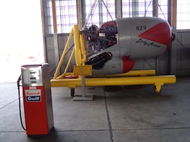 An old gasoline pump with the brand 'Good Gulf' stands next to a disassembled aircraft engine display labeled 'Lady Annie'. The engine is mounted on a yellow stand inside an industrial hangar with large windows.