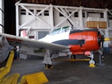 A vintage military aircraft is displayed inside a hangar. The airplane has a predominantly white body with distinctive red and black markings, featuring a red nose and a yellow propeller tip. The hangar shows structural beams and windows in the background, while a sign on the ground provides information about the aircraft.